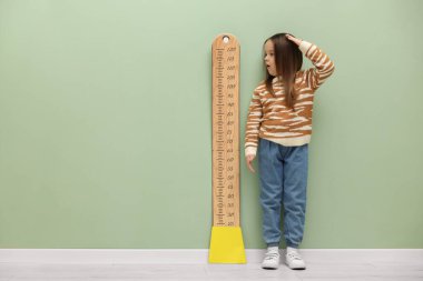 Açık yeşil duvarın yanında boyunu çizelgeyle ölçen küçük bir kız. Metin için boşluk