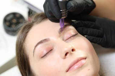 Güzellik uzmanı, içeride genç bir kadına kalıcı kaş makyajı yapıyor.