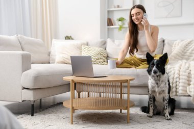 Kanepede akıllı telefonuyla konuşan gülümseyen kadın ve evde sevimli köpeği.