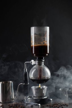 Vakumlu kahve makinesinde aromatik kahve demlemek, laboratuvar bardağı ve fasulye gri masada koyu arkaplanda dumanla kaplanmak, yakın plan