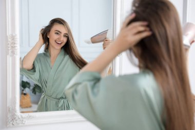 Bornozlu gülümseyen kadın banyoda aynanın yanında saç kurutma makinesiyle saçını kurutuyor.