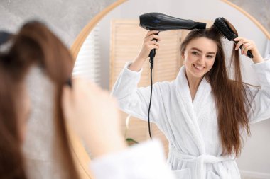 Bornozlu gülümseyen kadın saçını saç kurutma makinesiyle şekillendiriyor ve banyoda aynanın yanında tarıyor.