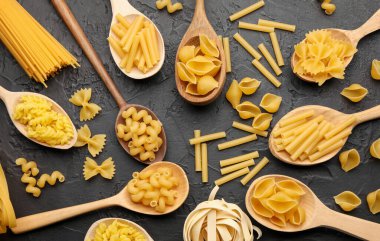 Tahta kaşıklardaki farklı çiğ makarna türleri siyah masada, düz yatıyordu.