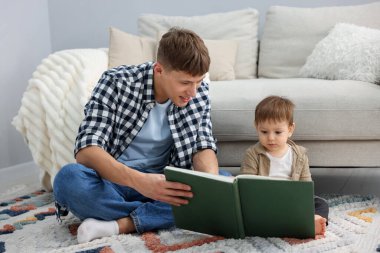 Gülümseyen erkek dadı evde yerde yatan şirin çocuğa kitap okuyor.