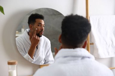 Handsome man applying lotion on his face near mirror in bathroom