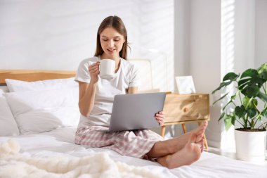 Elinde kahve olan bir kadın evde dizüstü bilgisayarla çalışıyor.