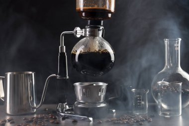 Vakumlu kahve makinesinde aromatik kahve demlemek, laboratuvar bardağı ve fasulye gri masada koyu arkaplanda dumanla kaplanmak, yakın plan