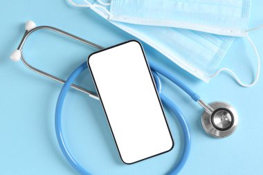 Sağlığınıza. Akıllı telefon, steteskop ve açık mavi arka planda tıbbi maskeler.