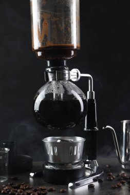 Vakumlu kahve makinesinde aromatik kahve demlemek, laboratuvar bardağı ve fasulye gri masada koyu arkaplanda dumanla kaplanmak, yakın plan