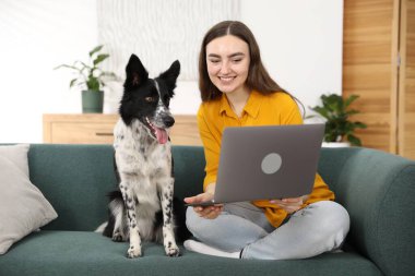 Evdeki kanepede dizüstü bilgisayarı ve sevimli köpeğiyle gülümseyen kadın.