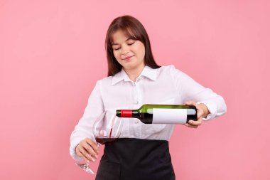 Profesyonel şarap garsonu pembe arka planda kırmızı şarabı bardağa dolduruyor.