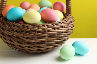 Güzel dekore edilmiş Paskalya yumurtaları hasır sepetin içinde beyaz ahşap masada sarı arka planda, yakın planda.