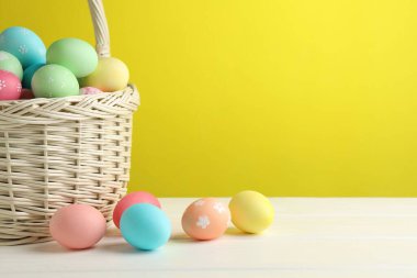 Hasır sepetteki güzel Paskalya yumurtaları beyaz ahşap masada, sarı arka planda, yakın plan. Metin için boşluk