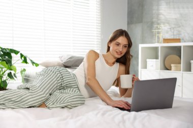 Elinde kahve olan bir kadın evde dizüstü bilgisayarla çalışıyor.