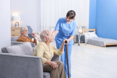 Bakım işçisi yaşlı kadına bastonuyla yardım ediyor huzurevinde koltuktan kalkması için.