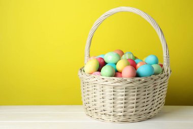 Güzel dekore edilmiş Paskalya yumurtaları hasır sepetin içinde beyaz ahşap masa üzerinde sarı arkaplan, metin için boşluk