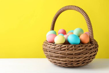 Güzel dekore edilmiş Paskalya yumurtaları hasır sepetin içinde beyaz ahşap masa üzerinde sarı arkaplan, metin için boşluk