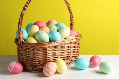 Güzel dekore edilmiş Paskalya yumurtaları hasır sepetin içinde beyaz ahşap masada sarı arka planda, yakın planda.