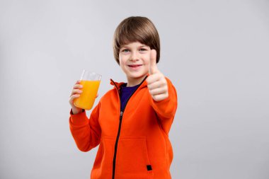 Bir bardak taze portakal suyu olan çocuk açık gri arka planda baş parmağını gösteriyor.
