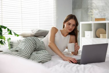 Elinde kahve olan bir kadın evde dizüstü bilgisayarla çalışıyor.