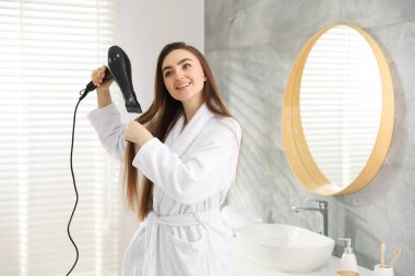 Bornozlu gülümseyen kadın banyoda saçını saç kurutma makinesiyle kurutuyor. Metin için boşluk