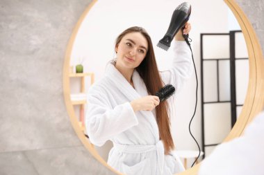 Bornozlu güzel bir kadın saçını saç kurutma makinesiyle şekillendiriyor ve banyoda aynanın yanında tarıyor. Metin için boşluk
