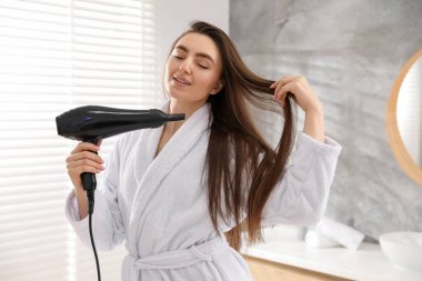 Bornozlu gülümseyen kadın saçını banyoda saç kurutma makinesiyle kurutuyor.