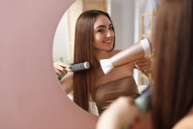 Gülümseyen kadın saçını saç kurutma makinesiyle şekillendiriyor ve içeride aynanın yanında fırçalıyor. Metin için boşluk