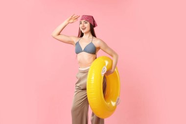 Pembe arka planda şişirilebilir yüzüklü mayo ve pantolon giymiş şık bir kadın.