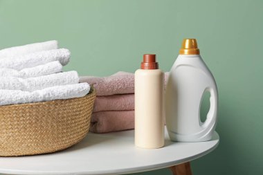 İçerideki yeşil duvarın yanındaki masada havlu ve şişe deterjan dolu çamaşır sepeti.