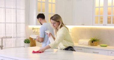 Kadın beyaz mermer masayı temizlerken adam mutfakta tabak siliyor.