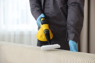 Haşere kontrol işçisi kanepeyi temizliyor. Kapalı alanda buhar temizleyicisi var.