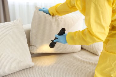 Koruyucu giysili haşere kontrol görevlisi. Kanepeyi inceleyip, el feneriyle kapatıyor.