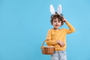 Paskalya kutlaması. Saçbantlı, tavşan kulaklı ve açık mavi arka planda boyalı yumurta dolu sepeti olan gülümseyen küçük bir çocuk. Metin için boşluk