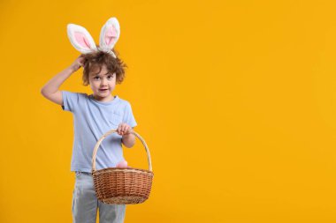 Paskalya kutlaması. Saç bantlı, tavşan kulaklı ve sarı arka planda boyalı yumurtalarla dolu sepetli şirin bir çocuk. Metin için boşluk