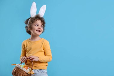 Paskalya kutlaması. Saçbantlı, tavşan kulaklı ve açık mavi arka planda boyalı yumurta dolu sepeti olan gülümseyen küçük bir çocuk. Metin için boşluk