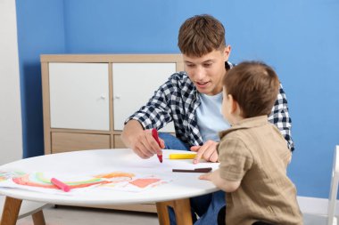 Gülümseyen erkek dadı ve beyaz masaya resim çizen sevimli küçük çocuk.