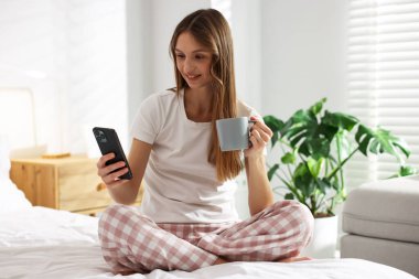 Evdeki yatakta bir fincan kahve ve akıllı telefonla güzel bir kadın.