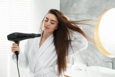 Bornozlu güzel bir kadın banyoda saçını saç kurutma makinesiyle kurutuyor.