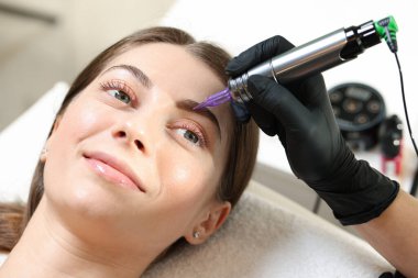 Güzellik uzmanı, kuaförde genç bir bayana kalıcı göz kalemi makyajı yapıyor.