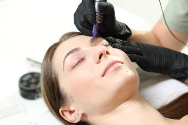 Güzellik uzmanı, içeride genç bir kadına kalıcı kaş makyajı yapıyor.