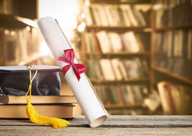 Diploma, mezuniyet şapkası ve kütüphanedeki tahta masanın üzerinde kitaplar. Kırmızı kurdeleyle kaydır