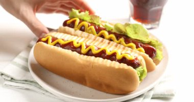 Beyaz ahşap masada ketçaplı, hardallı ve marullu lezzetli sosisli sandviç yiyen bir kadın.