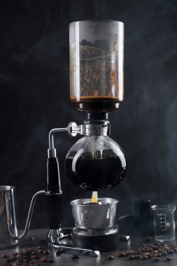 Vakumlu kahve makinesinde aromatik kahve demlemek, laboratuvar bardağı ve fasulye gri masada koyu arkaplanda dumanla kaplanmak, yakın plan