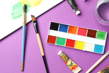 Suluboya boya, fırça, boya, bir bardak su ve mor zemin üzerinde tüpler olan palet.