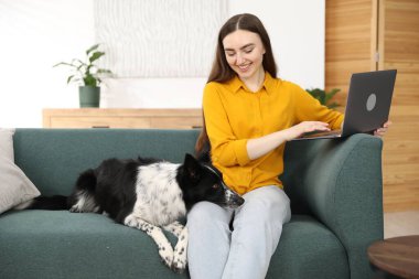 Evdeki kanepede dizüstü bilgisayarı ve sevimli köpeğiyle gülümseyen kadın.