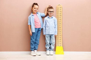 Küçük çocuk ve kız bej duvarın yanında boylarını grafikle ölçüyorlar.