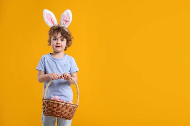 Paskalya kutlaması. Saç bantlı, tavşan kulaklı ve sarı arka planda boyalı yumurtalarla dolu sepetli şirin bir çocuk. Metin için boşluk