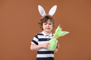 Paskalya kutlaması. Saç bandı takmış, tavşan kulaklı, kahverengi arka planda hediye tutan sevimli bir çocuk portresi.