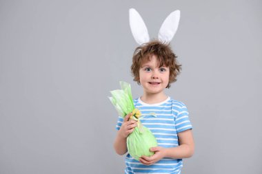 Paskalya kutlaması. Saçbandı takmış, tavşan kulaklı gülümseyen çocuğun portresi gri arka planda hediye tutuyor, mesaj için yer var.